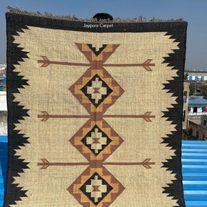 May include: A rectangular rug with a beige central field featuring brown arrow and diamond patterns. The rug has a black border with a zigzag design and a fringed edge. The text "Jaypore Carpet" is visible.