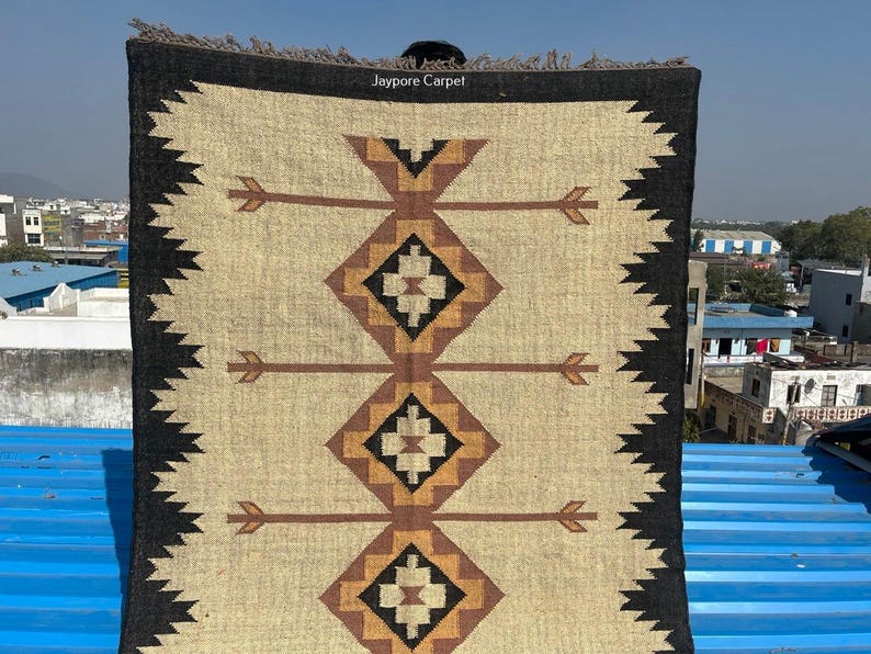 May include: A rectangular rug with a beige central field featuring brown arrow and diamond patterns. The rug has a black border with a zigzag design and a fringed edge. The text "Jaypore Carpet" is visible.