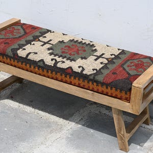 May include: Wooden bench with a rectangular seat featuring a patterned fabric in red, green, black, and beige. The bench has a light brown wooden frame and angled legs, suitable for indoor or outdoor use.