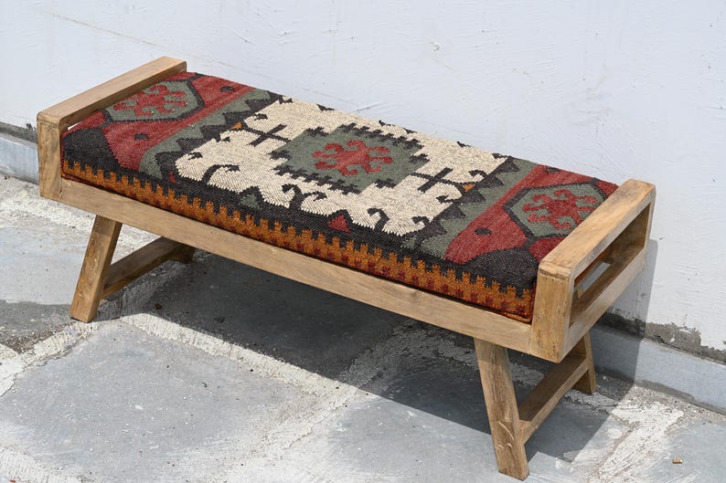 May include: Wooden bench with a rectangular seat featuring a patterned fabric in red, green, black, and beige. The bench has a light brown wooden frame and angled legs, suitable for indoor or outdoor use.
