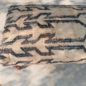 Op de afbeelding: Een rechthoekige poef van jute met een herhalend zwart geometrisch patroon op een natuurlijke beige achtergrond. De poef staat op vier kleine, afgeronde houten poten en biedt een mix van textuur en design. Ongeveer 120 cm lang.