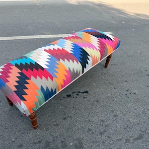May include: A rectangular bench with a colorful, geometric patterned top. The pattern includes zigzags in shades of red, blue, orange, and white. The bench has four wooden legs.