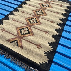 May include: Rectangular beige rug with a black border and fringe. The rug features a repeating pattern of brown diamonds and arrows. The text "Jaypore Carpet" is visible on the rug.