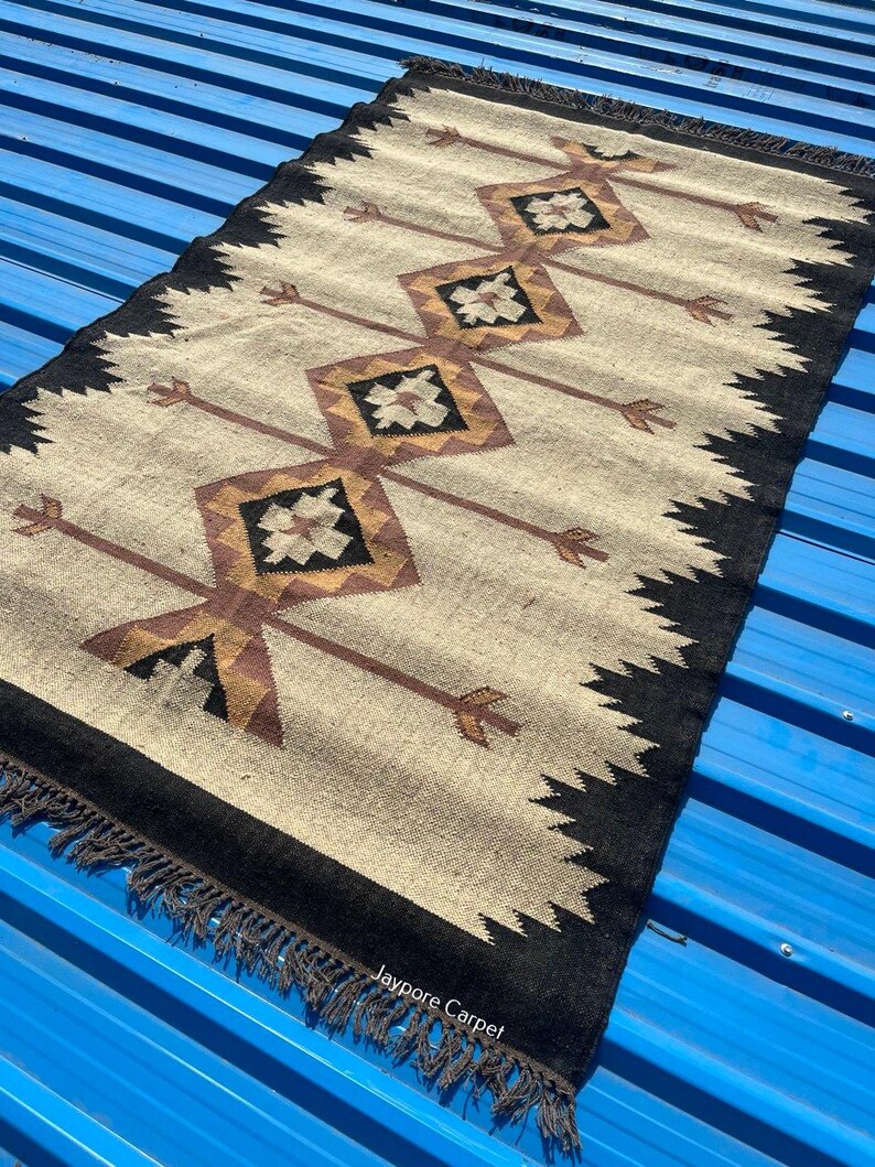 May include: Rectangular beige rug with a black border and fringe. The rug features a repeating pattern of brown diamonds and arrows. The text "Jaypore Carpet" is visible on the rug.