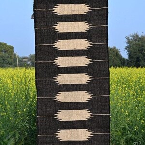 May include: A black and beige woven rug with a geometric pattern of horizontal beige shapes against a black background. The rug is hanging outdoors, with the text "Jaypore Carpet" visible at the top. The rug is displayed against a backdrop of yellow flowers.