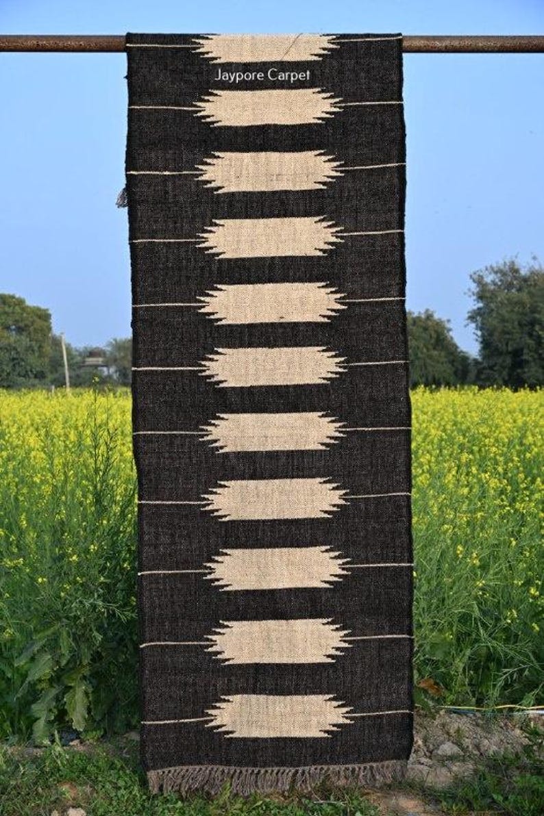 May include: A black and beige woven rug with a geometric pattern of horizontal beige shapes against a black background. The rug is hanging outdoors, with the text "Jaypore Carpet" visible at the top. The rug is displayed against a backdrop of yellow flowers.