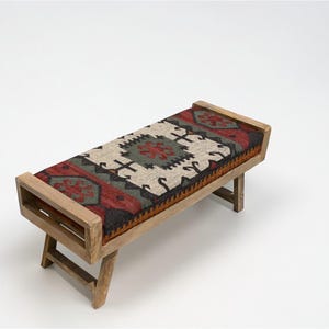 May include: A wooden bench with a rectangular seat featuring a colorful, patterned rug design in red, green, black, and cream. The bench has a light brown wooden frame and angled legs, suitable for indoor use.