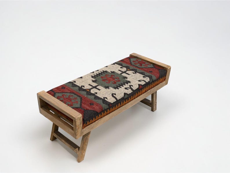 May include: A wooden bench with a rectangular seat featuring a colorful, patterned rug design in red, green, black, and cream. The bench has a light brown wooden frame and angled legs, suitable for indoor use.