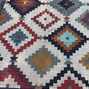 May include: A woven rug with a geometric diamond pattern in shades of red, blue, brown, and gray on a cream background. The rug has a fringed edge and a textured appearance, suitable for home decor.