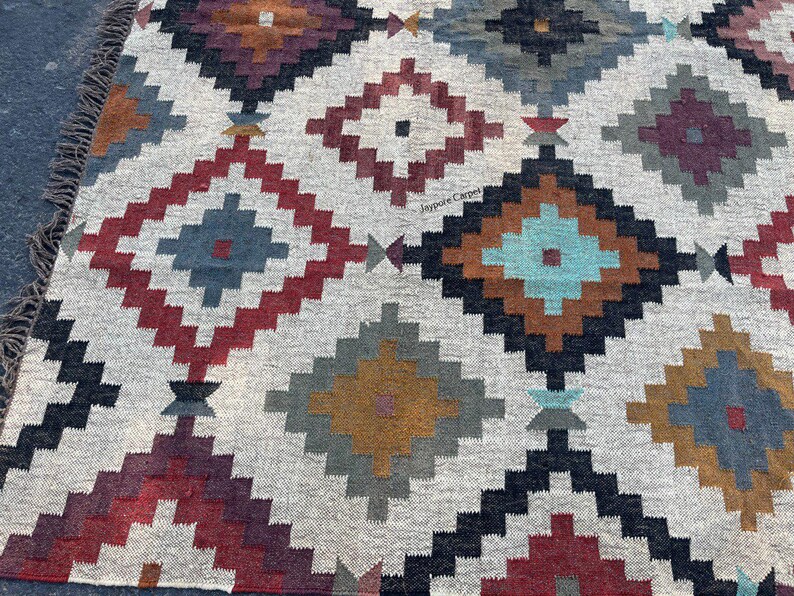 May include: A woven rug with a geometric diamond pattern in shades of red, blue, brown, and gray on a cream background. The rug has a fringed edge and a textured appearance, suitable for home decor.