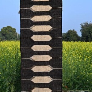 May include: A black and beige rectangular rug with a geometric design. The rug has alternating bands of black and beige, with a fringe at the bottom. It's displayed outdoors against yellow flowers and a blue sky. The text "Jaypore Carpet" is visible.