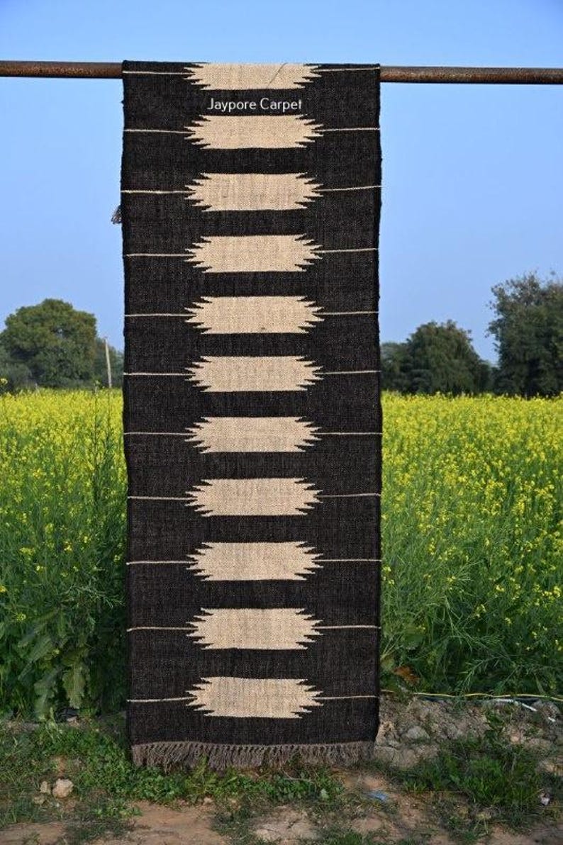 May include: A black and beige rectangular rug with a geometric design. The rug has alternating bands of black and beige, with a fringe at the bottom. It's displayed outdoors against yellow flowers and a blue sky. The text "Jaypore Carpet" is visible.