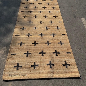 May include: A rectangular jute rug featuring a repeating pattern of black plus signs. The rug has a natural tan colour and a woven texture. The text "Jaypore Carpet" is visible on the rug.