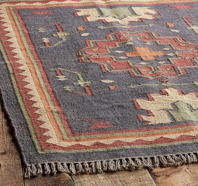 Pode incluir: Um tapete retangular com um padr&atilde;o geom&eacute;trico em tons de cinza, vermelho, laranja, verde e bege. O tapete tem uma borda com franjas e uma borda com um design de tri&acirc;ngulo repetido. O tapete &eacute; feito de um material tecido.