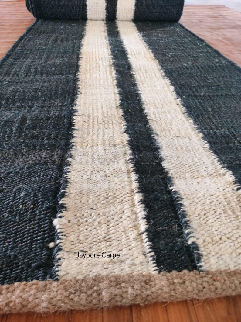 May include: A rolled-up, rectangular rug with a dark blue-gray background and two wide, off-white stripes running lengthwise. The rug has a textured appearance and a brown border. The text "Jaypore Carpet" is visible on the rug.