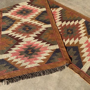 May include: Two matching woven table runners with a geometric pattern in shades of brown, pink, cream, and navy blue. The runners have a fringed edge and are suitable for a dining table or entryway.