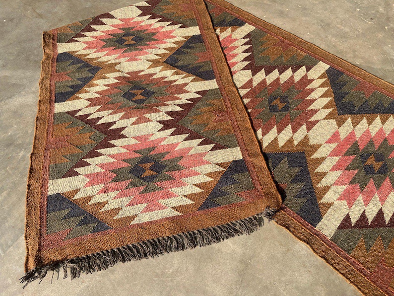 May include: Two matching woven table runners with a geometric pattern in shades of brown, pink, cream, and navy blue. The runners have a fringed edge and are suitable for a dining table or entryway.