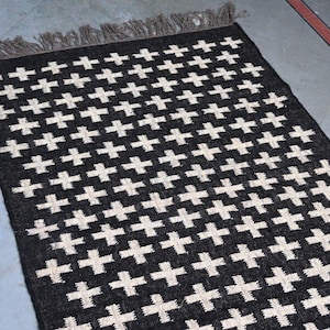Puede incluir: Una alfombra rectangular negra con un patrón repetido de cruces blancas. La alfombra tiene un borde con flecos en un color neutro. La alfombra está hecha de un material tejido y es adecuada para uso en interiores.