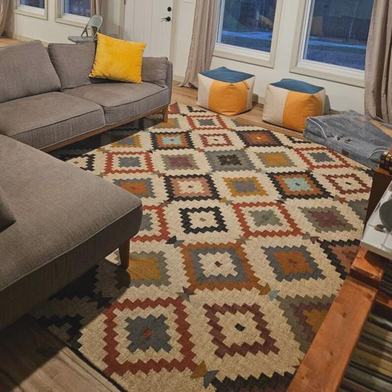 May include: A large area rug with a geometric pattern in shades of beige, brown, orange, blue, and gray. The rug is placed in a living room setting, with a gray sofa and yellow pillow visible.