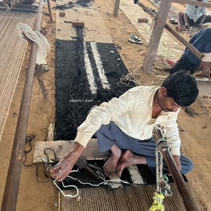 May include: A rug in progress, featuring a black and white striped design. The rug is being woven on a wooden loom, with a craftsman working on the details. The image shows the process of handmade carpet creation, with the text "Jaypore Carpet" visible.