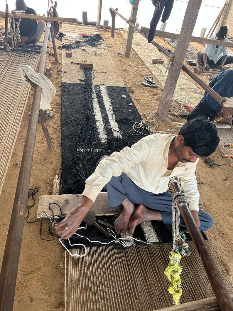 May include: A rug in progress, featuring a black and white striped design. The rug is being woven on a wooden loom, with a craftsman working on the details. The image shows the process of handmade carpet creation, with the text "Jaypore Carpet" visible.