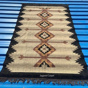 May include: Rectangular beige rug with a geometric pattern in brown, black, and tan. The rug has a black border with fringe and arrow-like designs. The text "Jaypore Carpet" is visible.