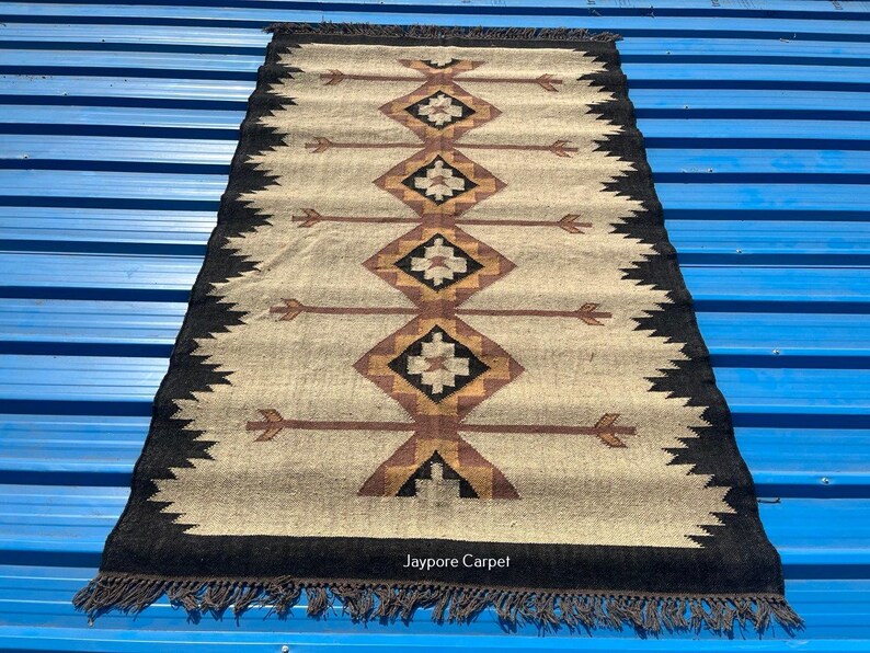 May include: Rectangular beige rug with a geometric pattern in brown, black, and tan. The rug has a black border with fringe and arrow-like designs. The text "Jaypore Carpet" is visible.
