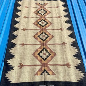 May include: Rectangular beige rug with a black border and geometric patterns in brown and black. The rug features diamond shapes and arrow-like designs. The edges have a fringed trim. The rug is made of a woven material.