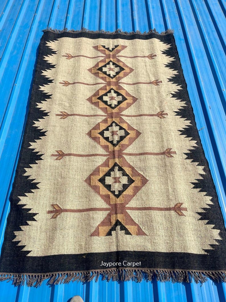 May include: Rectangular beige rug with a black border and geometric patterns in brown and black. The rug features diamond shapes and arrow-like designs. The edges have a fringed trim. The rug is made of a woven material.