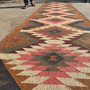 May include: A long, rectangular jute rug with a geometric pattern in shades of brown, pink, cream, and navy blue. The rug has a textured surface and a brown border. The rug is suitable for a hallway or entryway.