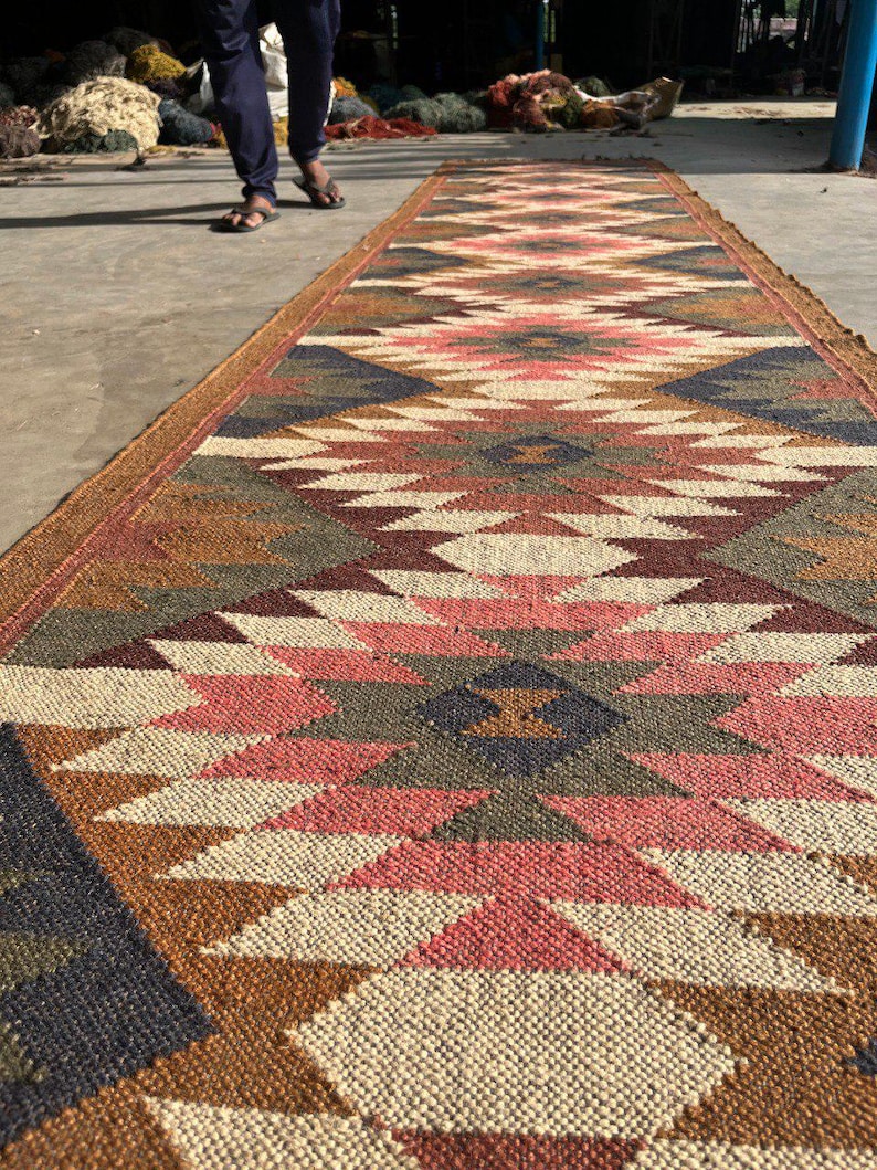 May include: A long, rectangular jute rug with a geometric pattern in shades of brown, pink, cream, and navy blue. The rug has a textured surface and a brown border. The rug is suitable for a hallway or entryway.