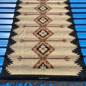May include: Rectangular beige rug with a geometric pattern in brown, black, and tan. The design features diamond shapes and arrow motifs. The rug has a black border with a fringe.
