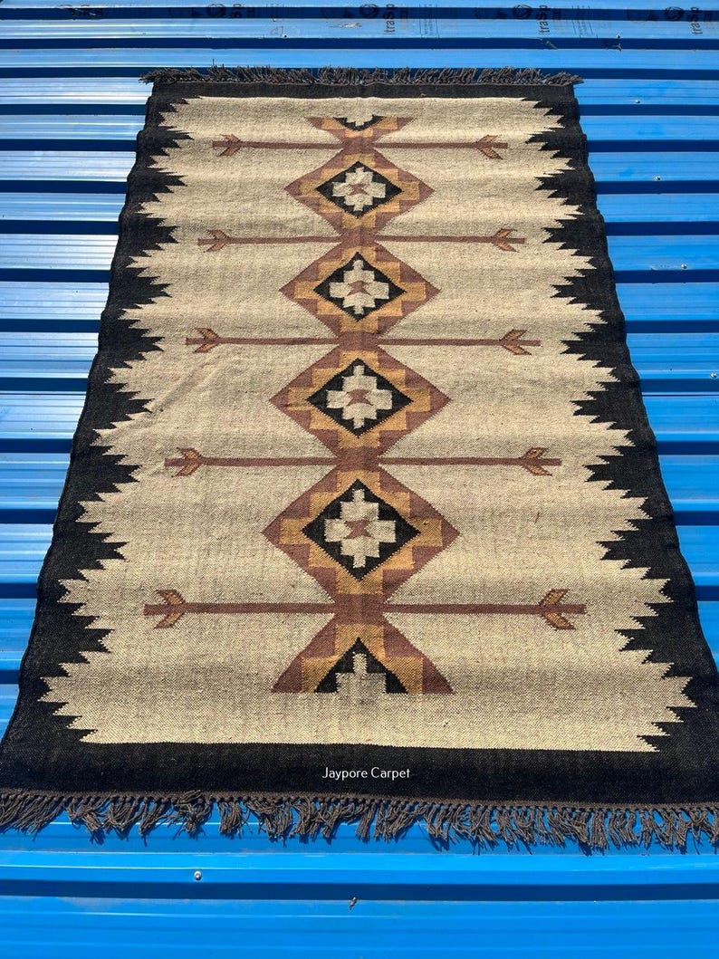 May include: Rectangular beige rug with a geometric pattern in brown, black, and tan. The design features diamond shapes and arrow motifs. The rug has a black border with a fringe.