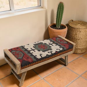 May include: A wooden bench with a rectangular seat featuring a colorful, patterned fabric in red, black, green, and cream. The bench has a light-colored wooden frame and legs. A cactus in a terracotta pot and a woven basket are nearby.