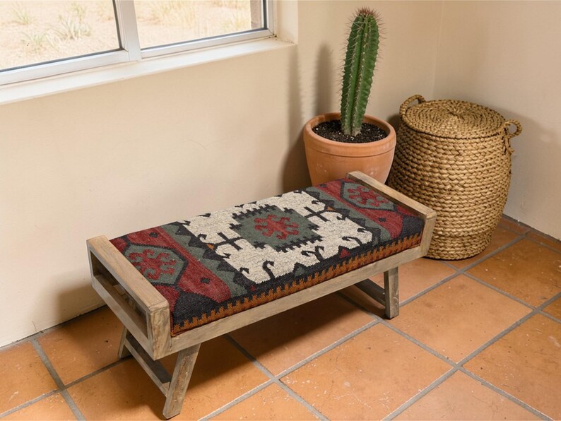 May include: A wooden bench with a rectangular seat featuring a colorful, patterned fabric in red, black, green, and cream. The bench has a light-colored wooden frame and legs. A cactus in a terracotta pot and a woven basket are nearby.