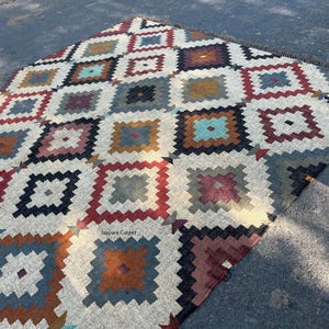 May include: A colorful, geometric patterned rug with a repeating square design. The rug features a cream-colored background with squares in shades of red, blue, brown, and teal. The rug is made of a woven material and has a fringe edge.
