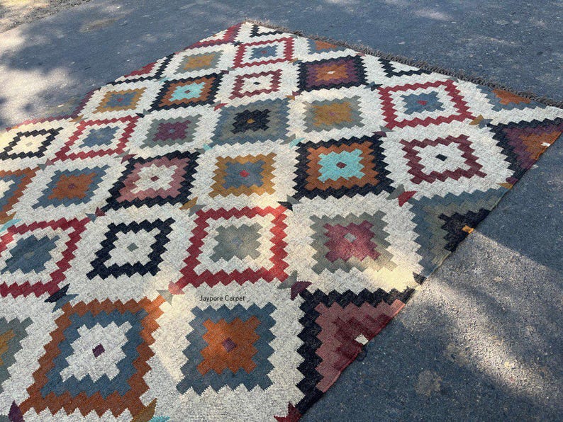 May include: A colorful, geometric patterned rug with a repeating square design. The rug features a cream-colored background with squares in shades of red, blue, brown, and teal. The rug is made of a woven material and has a fringe edge.