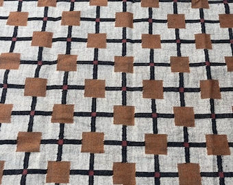 Könnte beinhalten: Gewebter Textil mit sich wiederholendem geometrischen Muster. Das Design zeigt braune Quadrate, schwarze Linien und kleine rote Akzente auf hellbeigem Hintergrund. Der Rand ist einfarbig rot. Der Textil hat einen Fransenrand.
