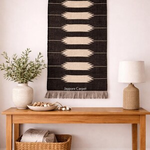 May include: A black and cream-coloured woven wall hanging with a geometric design hangs above a wooden console table. The table features a lamp, a vase with greenery, and a woven basket. The text "Jaypore Carpet" is visible on the wall hanging.