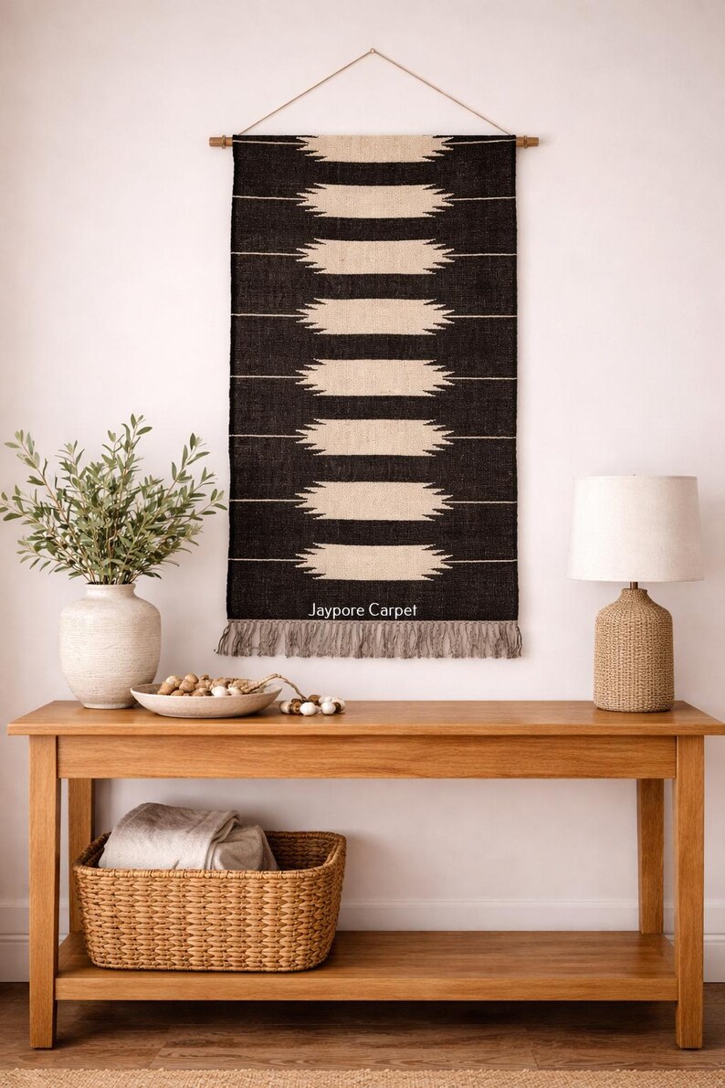 May include: A black and cream-coloured woven wall hanging with a geometric design hangs above a wooden console table. The table features a lamp, a vase with greenery, and a woven basket. The text "Jaypore Carpet" is visible on the wall hanging.