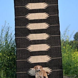 May include: A black and beige patterned rug with the text "Jaypore Carpet" is displayed outdoors. A small tan puppy stands in front of the rug. The rug has a repeating geometric design.