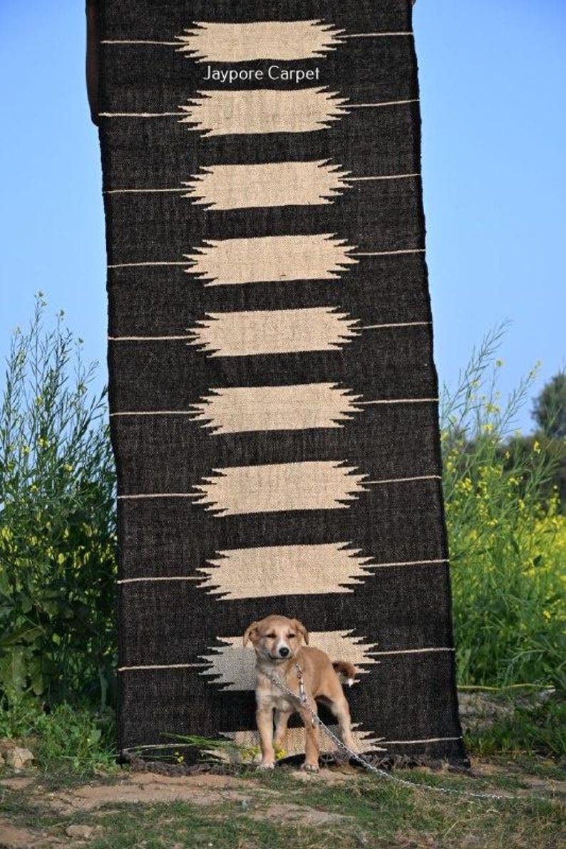 May include: A black and beige patterned rug with the text "Jaypore Carpet" is displayed outdoors. A small tan puppy stands in front of the rug. The rug has a repeating geometric design.