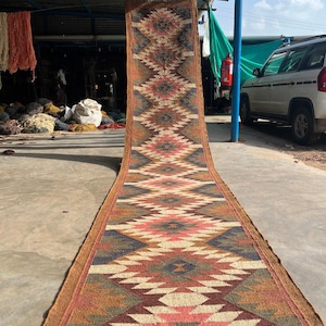 May include: A long, rectangular rug with a geometric pattern in shades of red, pink, brown, beige, and blue. The rug has a brown border and is displayed outdoors. The rug is made of woven material.