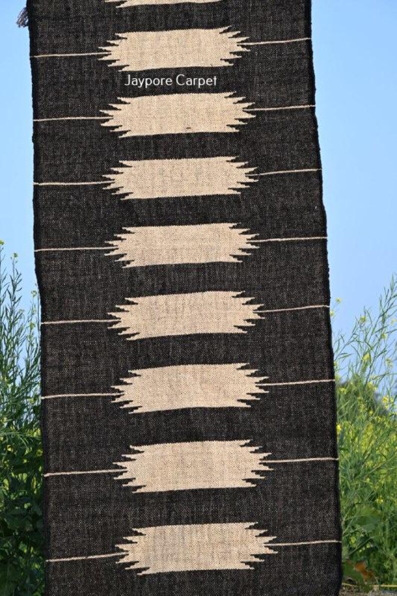 May include: A black and beige woven rug with a geometric pattern. The rug features horizontal beige stripes with jagged edges against a black background. The text "Jaypore Carpet" is visible.