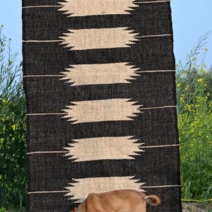 May include: A black and beige woven rug with a geometric pattern. The rug features horizontal beige shapes against a black backdrop. The text "Jaypore Carpet" is visible. A small brown dog is sniffing the ground near the rug.
