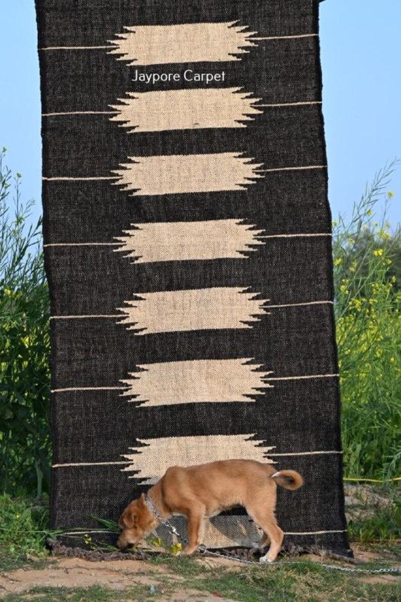 May include: A black and beige woven rug with a geometric pattern. The rug features horizontal beige shapes against a black backdrop. The text "Jaypore Carpet" is visible. A small brown dog is sniffing the ground near the rug.