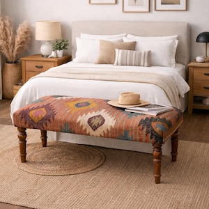 Può includere: Interno di camera da letto con letto, panca fantasia e quadri incorniciati. La panca ha un colorato motivo geometrico. Un cappello di paglia e libri sono sulla panca. La stanza ha una palette di colori neutri con mobili in legno e un tappeto in juta. La panca è lunga circa 120 cm.