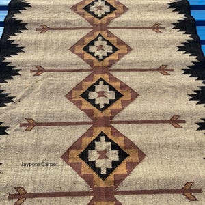 May include: A rectangular beige rug with a geometric pattern. The rug features repeating diamond shapes in brown, black, and gold, with arrow-like designs. The edges are bordered with a black zigzag pattern. The text "Jaypore Carpet" is visible.