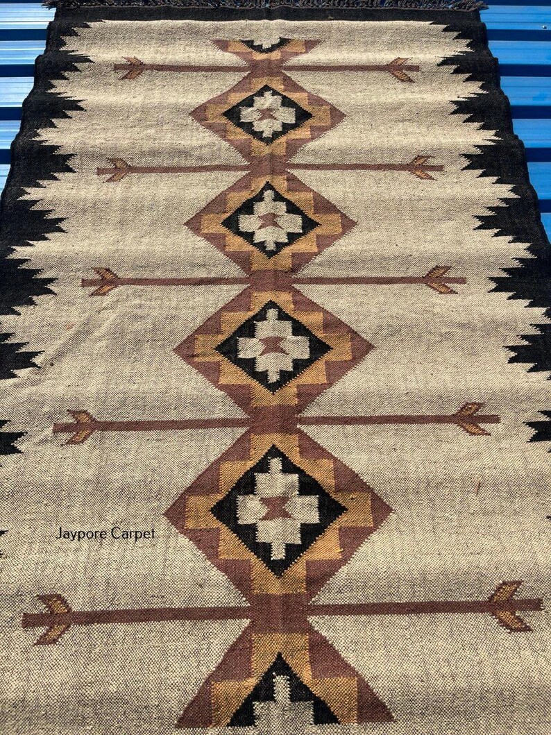 May include: A rectangular beige rug with a geometric pattern. The rug features repeating diamond shapes in brown, black, and gold, with arrow-like designs. The edges are bordered with a black zigzag pattern. The text "Jaypore Carpet" is visible.