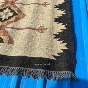 May include: A beige and black woven rug with a geometric pattern. The rug features diamond shapes in brown, black, and tan, with a black border and fringe. The text "Jaypore Carpet" is visible.
